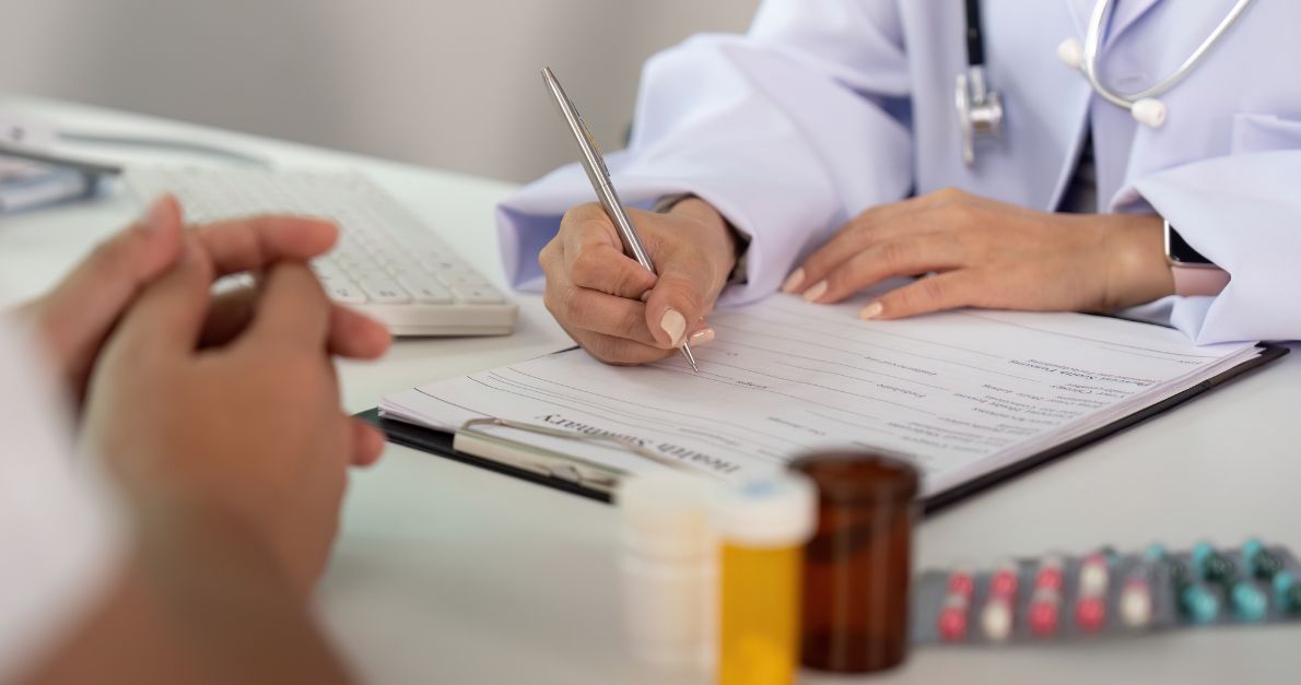 Clinician in white coat completing a medical form on a clipboard, with patient hands, keyboard, pill bottles and blister packs on the desk.