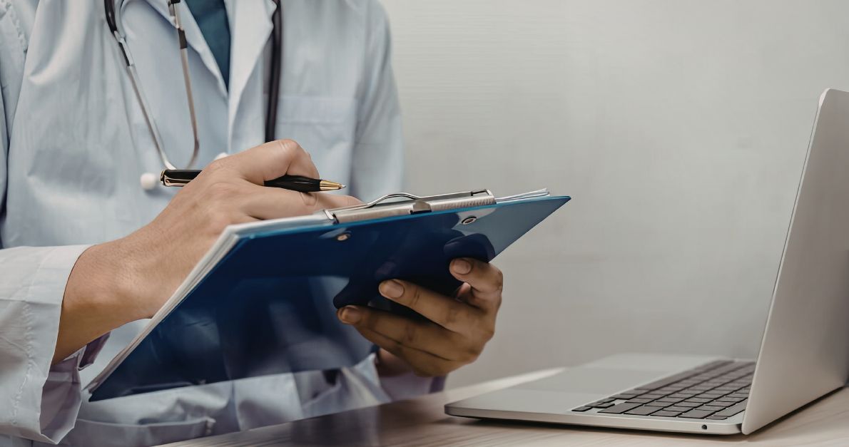 Doctor writing on a clipboard beside a laptop, representing clinical trial documentation, SOP management, and research oversight.